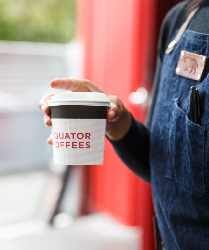 Person holding an Equator Coffees coffee cup, wearing a blue apron | Equator Coffees